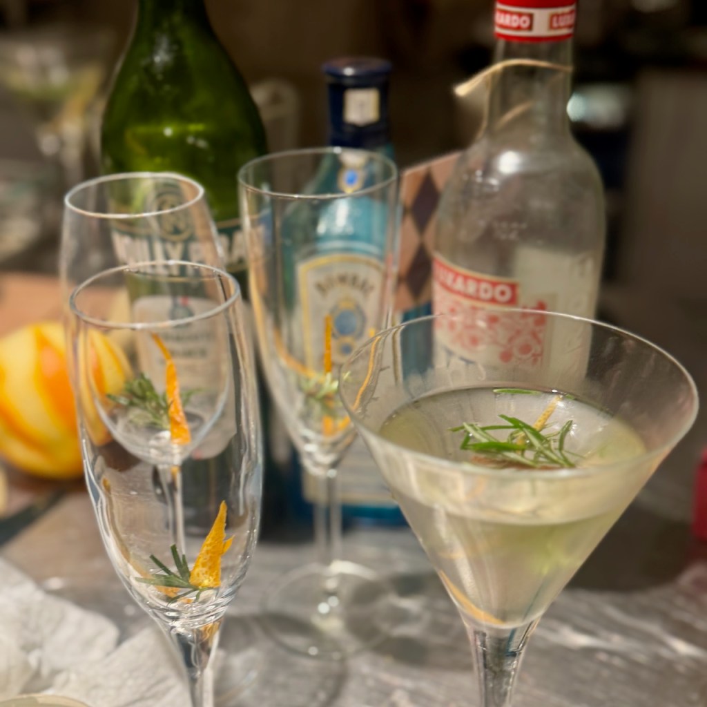 A white negroni cocktail (gin, white vermouth, bitter bianco) served in a cocktail glass garnished with a twist of orange and a sprig of rosemary.
